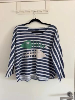 Randig stickad tröja med fiskmotiv - Blåvit randig stickad tröja med ett stort fiskbensmotiv i grönt, vitt och beige på framsidan. Tröjan har rund halsringning och trekvartsärmar. Materialet ser mjukt och bekvämt ut, perfekt för en avslappnad stil. Sparsamt använd