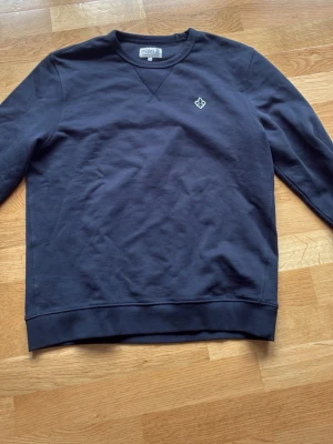 Mörkblå sweatshirt från Morris - Klassisk mörkblå sweatshirt från Morris med rund halsringning och diskret logga broderad på bröstet. Tröjan har ribbade muddar vid ärmslut och nederkant samt en mjuk bomullskänsla. Perfekt för en avslappnad och stilren look. Storlek M och passar perfekt för folk mellan 168-180 cm
