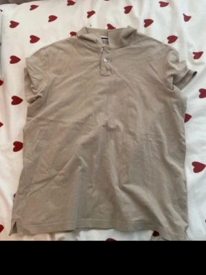 Beige henley från Pier One - Säljer en beige henley från Pier One med korta ärmar och knappar vid halsen. Skjortan är i mjuk bomull och har en avslappnad passform. Perfekt för dig som gillar en enkel och stilren look.