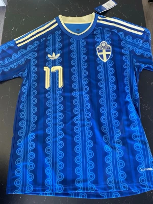Blå Sverige bortatröja 2026 - Säljer en blå Sverige fotbollströja med retroinspirerat mönster, gula detaljer och nummer 17 samt namnet Gyökeres på ryggen. Tröjan har korta ärmar, rund hals och broderat landslagsemblem på bröstet. Tillverkad i lätt och ventilerande material.