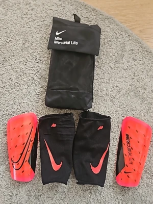 Nike Mercurial benskydd med sleeves - Säljer ett par Nike Mercurial Lite benskydd i neonrött och svart med tillhörande svarta sleeves och förvaringspåse. Benskydden har ventilerande hål och mjuk vit baksida för extra komfort. Perfekta för fotbollsträning eller match. Helt nya, använda max 2 gånger. Säljer de eftersom jag har andra benskydd. Kom med frågor.