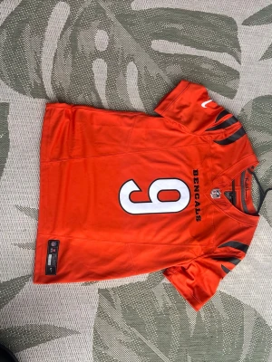 Cincinnati Bengals matchtröja orange - Snygg orange matchtröja med nummer 9 och namnet Burrow på ryggen. Tröjan har svarta detaljer på axlarna och är kortärmad med sportig passform. Perfekt för dig som vill visa ditt stöd för Bengals på matchen eller träningen.
