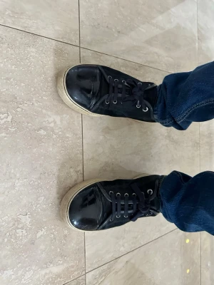 Lanvin - Säljer ett par svarta Lanvin med glansig tå och sidor i mockaliknande material. Skorna har svarta skosnören, metallfärgade öljetter och vit sula med diskret mönster. Klassisk design som funkar till jeans och streetwear.