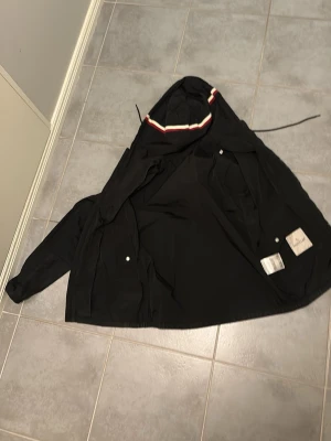 Svart vindjacka med huva - Snygg svart vindjacka med huva och dragsko. Jackan har vita och röda detaljer på huvan samt två framfickor med tryckknappar. Materialet är tunt och smidigt, perfekt för blåsiga dagar. Klassisk och stilren design. Den är i storlek 1 i monclear storlek