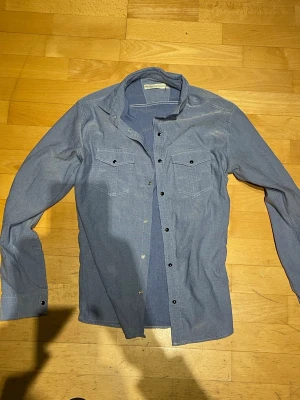 Blå långärmad skjorta med knappar - Snygg blå skjorta med lång ärm och tryckknappar framtill. Två bröstfickor med lock och knappdetaljer ger en klassisk look. Perfekt till jeans eller chinos för en avslappnad stil.