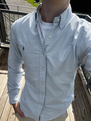 Blåvit randig skjorta från Ralph Lauren - Säljer en klassisk blåvit randig skjorta från Ralph Lauren med broderad logga på bröstet. Skjortan har lång ärm, knappar framtill och button-down krage. Perfekt till jeans eller chinos för en clean och stilren look.