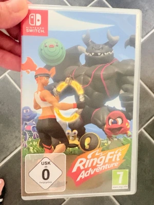 Ring Fit Adventure till Switch - Ring Fit Adventure till Nintendo Switch är ett träningsspel där du använder en ringformad kontroll och Joy-Con för att utföra olika övningar och utmana en stor svart muskelboss. Färgglad grafik och äventyrstema gör träningen roligare.