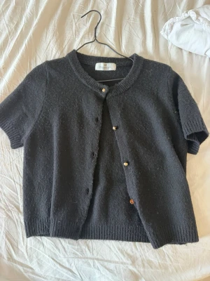 Mörkgrå kortärmad kofta från H&M - Kortärmad kofta i mörkgrått från H&M med rund halsringning och ribbade kanter. Koftan har knappar framtill och är gjord i ett mjukt stickat material. Perfekt att slänga över en topp för en chill look.