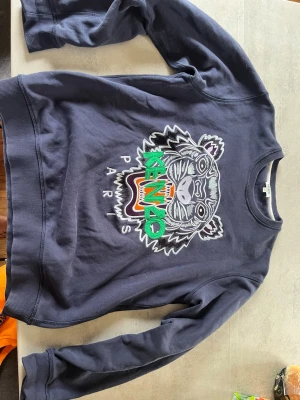 Mörkblå Kenzo sweatshirt med tiger - mörkblå sweatshirt från Kenzo med det ikoniska tigermotivet broderat framtill i grönt, orange och vitt. Rund halsringning och ribbade muddar vid ärmslut och nederkant. Perfekt för dig som vill sticka ut med en streetstyle-look.