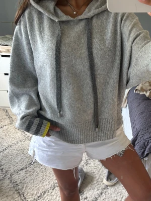 Grå hoodie  - Mysig grå hoodie med huva och långa snören framtill. Tröjan har breda ribbade muddar i grått och mörkgrått med en gul detalj längst ner på ärmarna. Oversized passform och mjukt material och tryck baktill