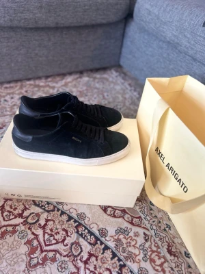  Axel Arigato skor - Stilrena svarta sneakers från Axel Arigato med ovandel i mocka och klassisk vit sula. Diskret logga i guld på sidan och svarta skosnören. Perfekta för dig som gillar minimalistisk och clean stil.