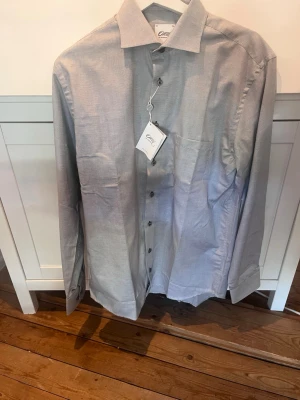 Oscar Jacobson skjorta  - Stilig ljusgrå/blå wrinkle free (regular fit) skjorta från Oscar Jacobson med klassisk krage och knappar framtill.  Skjortan har lång ärm och en bröstficka, perfekt för en stilren look. Endast testad! 