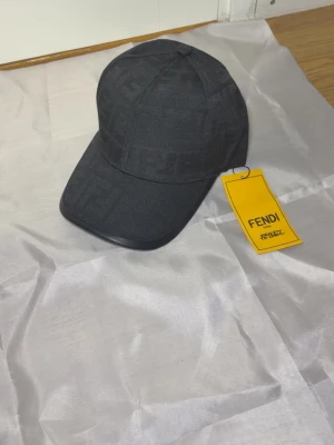 Svart keps från Fendi med logomönster - Snygg svart keps från Fendi med diskret FF-logomönster i tyget. Justerbar rem baktill i läder med Fendi-gravering. Klassisk böjd skärm och stilren design, perfekt för att lyfta din outfit.