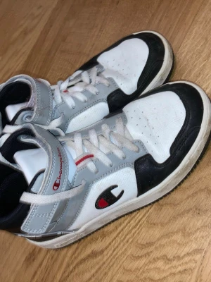 Champion High-Top Sneakers - Säljer ett par vita och svarta använda high-top Champion sneakers, med röda och grå detaljer. Skorna har vita snören och svart sula. Skorna är använda ett par fgr men funkar lika bra o använda flera ggr till.