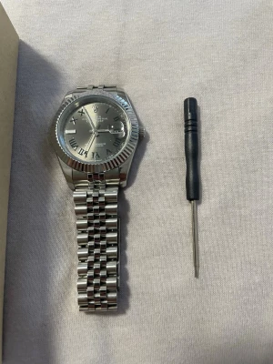 Silverfärgad klocka med romerska siffror - Stilren klocka i silver med metallarmband och romerska siffror på urtavlan. Klockan har en räfflad bezel och datumvisning. Medföljer verktyg för justering samt originalförpackning med skuminsats. OBS helt ny köpt, för att sälja vidare. Du får me allt på bindeln och du kan justera till hur du vill ha den , storlek på den.