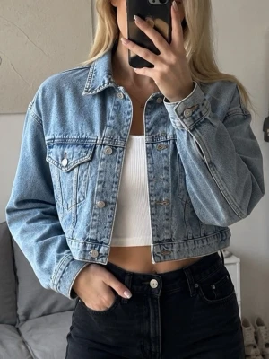 Ljusblå croppad jeansjacka - Trendig ljusblå croppad jeansjacka med klassisk krage och knappstängning framtill. Jackan har två bröstfickor med lock och silverfärgade knappar. Perfekt till ett par höga jeans eller kjol för en cool look.