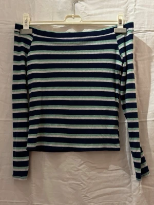 Randig långärmad off shoulder topp i blått och vitt - Säljer en långärmad off shoulder topp med marinblå och vita horisontella ränder. Toppen har en rak passform och är tillverkad i ett ribbat material som ger en skön stretchig känsla. Perfekt för dig som gillar en enkel men snygg stil. Ingen lapp kvar skulle säga att den passar xs-s