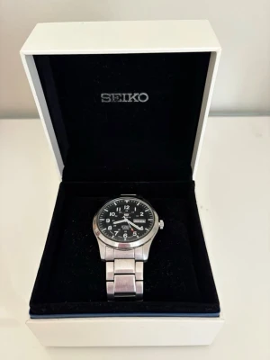 Seiko 5 Sports Automatisk Herr Klocka 100M - Stilren Seiko herrklocka med svart urtavla och tydliga vita siffror. Klockan har en robust länk i silverfärgat stål och dag- och datumvisning. Perfekt accessoar för dig som gillar klassisk och sportig stil.