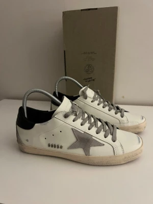 Vita Golden goose skor - Snygga vita sneakers med grå mockastjärna på sidan och grå skosnören. Skon har svart hälparti och en något smutsad sula för en trendig, använd look. Yttermaterialet är slätt läder och sulan är i gummi med fiskbensmönster.