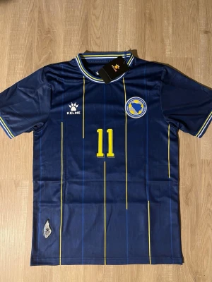 Blå Bosnien fotbollströja Dzeko #11 - Snygg mörkblå fotbollströja med gula detaljer och tunna ränder. Bröstet har Bosnien & Hercegovinas landslagsemblem och nummer 11. På ryggen står det Dzeko och nummer 11 i gult. Perfekt för match eller träning. HELT NY!