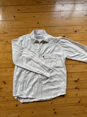 Vit jeansskjorta från Levi's - Vit långärmad jeansskjorta från Levi's med tryckknappar och bröstficka med klassisk röd Levi's-tag. Skjortan har en avslappnad passform och western-inspirerade sömmar. Perfekt att styla lager på lager eller bära som den är.