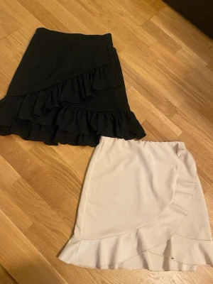 Två omlottkjolar med volang - Säljer två snygga korta omlottkjolar med volangdetaljer. En kjol är svart och den andra är beige, båda i ett mjukt och lätt material. Perfekta för sommarens alla tillfällen och ger en trendig look passar även stl s. 
