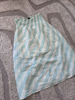 Randig långkjol från Lindex - Ljusblå och vit långkjol från Lindex med bred smock i midjan. Kjolen har ett diagonalt randigt mönster och är gjord i ett tunt, luftigt material som passar perfekt till varma dagar.