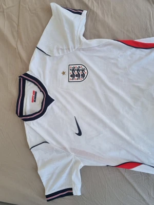 England Nike vm 2026 fotbollströja - Snygg vit England fotbollströja från Nike med broderad landslagssköld och guldstjärna på bröstet. Tröjan har marinblå och röda detaljer vid krage och ärmslut samt röda inslag på sidorna. Tillverkad i lätt och ventilerande material, perfekt för träning eller match.