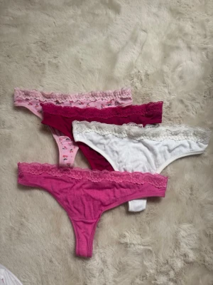 4-pack stringtrosor med spetskant - Säljer ett 4-pack stringtrosor i olika färger: rosa, vinröd, vit och ljusrosa med små hjärtan. Alla trosor har en ribbad struktur och en söt spetskant upptill. Perfekta för dig som gillar bekväma och snygga underkläder med lite extra detalj.