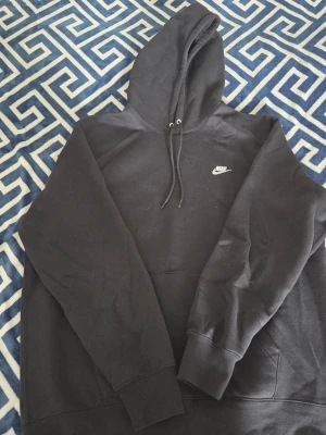Svart Nike hoodie med huva - Säljer en svart hoodie från Nike med klassisk huva och dragsnören. Hoodien har en stor magficka framtill och Nike-logga broderad i vitt på bröstet. Tillverkad i mjukt bomullsmaterial och har ribbade muddar vid ärmslut och nederkant.