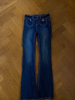 Mörkblå bootcut jeans med kontrastsömmar - Säljer ett par mörkblå bootcut jeans med vita kontrastsömmar och snygga detaljer på fickorna. Jeansen har låg midja, två framfickor med knappar och dekorativa bakfickor med broderade sömmar. Perfekta för en avslappnad och trendig look.