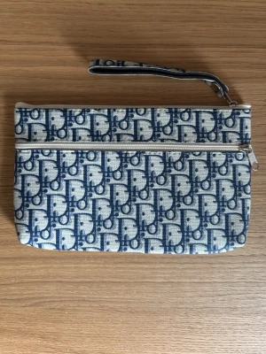 Blåvit kuvertväska med monogram mönster i blått/vittr - Snygg kuvertväska i tyg med blått och vitt monogrammönster. Väskan har en klassisk look och är trendig i både mönster och modell. Perfekt för att bära det viktigaste och ger en trendig touch till din outfit. Mobilen i går ej men ger en bild av hur stor väskan är. 