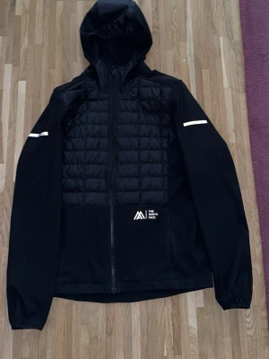 Svart vindjacka från The North Face - Snygg svart vindjacka från The North Face med quiltad front, hel dragkedja och huva. Jackan har vita detaljer på ärmarna och logga på bröstet. Tillverkad i ett lätt material som skyddar mot vind och har en sportig look.