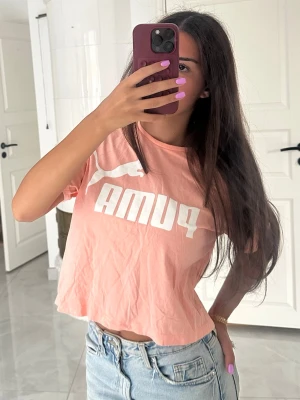 Ljusrosa croppad Puma t-shirt - Säljer en ljusrosa croppad t-shirt från Puma med vit logga framtill. T-shirten har korta ärmar och är tillverkad i mjuk bomull, perfekt för en avslappnad och trendig look.