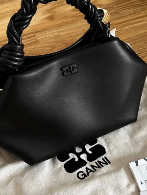 Ganni Svart handväska med flätat handtag - Snygg svart handväska i slätt läder med ett unikt flätat handtag och avtagbar axelrem med guldfärgade detaljer. Väskan har en modern, kantig form och diskret logga framtill. Perfekt för dig som vill ha en trendig accessoar med coola detaljer.