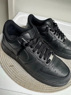 Nike Air Force 1 helsvarta sneakers - Klassiska Nike Air Force 1 sneakers i helsvart läder med svart sula och svarta skosnören.  på sidan och metallbricka med 'AF1' på snörningen. 
