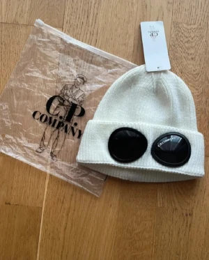 Vit mössa med goggles från C.P. Company - Vit ribbstickad mössa från C.P. Company med ikoniska svarta goggles på uppviket. Snygg och unik accessoar som sticker ut, perfekt för dig som gillar streetwear och vill ha något extra. Märkeslapp och originalpåse medföljer.