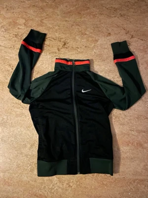  Nike zip tröja - Snygg svart och röd zip tröja från Nike med dragkedja framtill. Tröjan har hög krage, långa ärmar och röda detaljer vid krage och ärmslut. Perfekt för träning eller vardag .