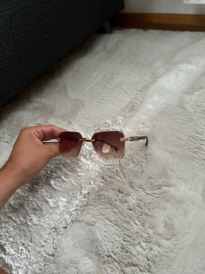 Bruna fyrkantiga solglasögon Givenchy - Snygga solglasögon från Givenchy med fyrkantiga, bruna glas och guldfärgade detaljer på skalmarna. Glasögonen har en modern design med genomskinliga näskuddar och bruna skalmar. Perfekt accessoar för att lyfta din stil.