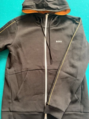 Svart zip hoodie från BOSS - Snygg svart hoodie från BOSS med dragkedja, huva med orange insida och svarta snören. Diskret BOSS-logga på bröstet och tunna gula detaljer längs ärmarna. Tillverkad i mjukt bomullsmaterial och har fickor framtill. Perfekt för en chill och stilren look.