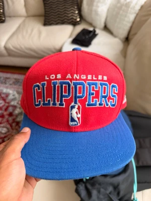 Los Angeles Clippers keps - Röd och blå snapback-keps med broderad Los Angeles Clippers-logga framtill och NBA-märke. Justerbar storlek med blå plastrem bak. Perfekt för basketfans som vill visa sitt lagstöd.