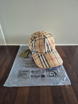  Burberry - Snygg beige keps från Burberry med det klassiska rutmönstret i svart, vitt och rött. Kepsen har böjd skärm och justerbar rem bak. Perfekt accessoar för en trendig och stilren look.