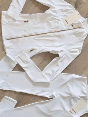 Vitt träningsset från Lululemon - Snyggt vitt träningsset från Lululemon med figurnära jacka och matchande tights. Jackan har dragkedja i guld, höga fickor med dragkedja och ståkrage. Materialet är stretchigt och följsamt, perfekt för träning eller yoga.