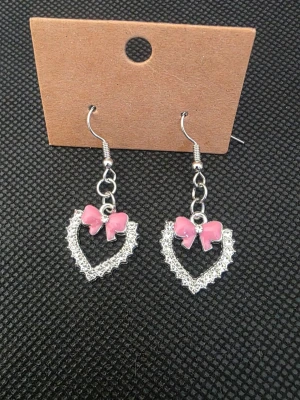 Hjärformade örhängen med rosa rosett - Söta örhängen i form av hjärtan med glittrande silverfärg och en liten rosa rosett upptill. Hängande modell med krok, perfekta för att ge en gullig touch till din stil. Materialet ser ut att vara metall med dekorativa detaljer.
