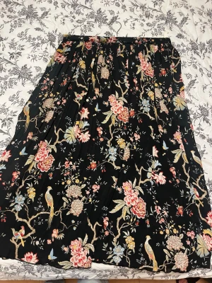 Svart blommig långkjol H&M x GP&J Baker - Säljer en svart långkjol från H&M x GP&J Baker med färgglatt blommönster. Kjolen har plisserad design och elastisk midja för skön passform. Perfekt för dig som gillar statement-plagg med unika detaljer.