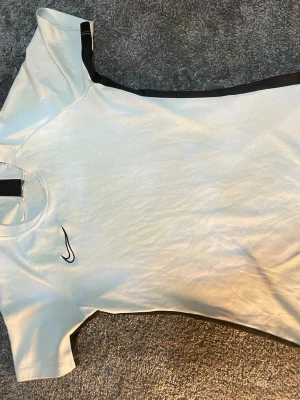 Vit Nike Dri-FIT träningströja XS - Säljer en vit Nike Dri-FIT träningströja i storlek XS. Tröjan har svarta detaljer längs axlarna och ett svart Nike-märke på bröstet. Tillverkad i ett lätt och ventilerande funktionsmaterial som passar perfekt till träning.