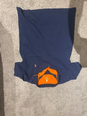 Marinblå piké från Polo Ralph Lauren - Snygg marinblå pikétröja från Polo Ralph Lauren med orange detaljer och klassisk logga broderad på bröstet. Tröjan har korta ärmar och en krage med orange insida som ger en cool kontrast. Perfekt för en avslappnad men stilren look.              Pris kan diskuteras.