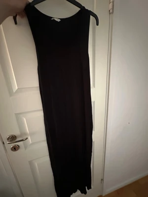 Svart ärmlös långklänning - En enkel och stilren svart långklänning med rund halsringning och ärmlös design. Klänningen har en rak passform och är tillverkad i ett mjukt, tunt material som faller snyggt längs kroppen. Perfekt för dig som gillar minimalistisk stil.