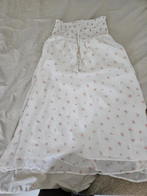 Långkjol - Söt vit långkjol från Gina Tricot med små rosa blommor och smock upptill. Kjolen har knytband med tofsar i midjan och är gjord i ett lätt, luftigt tyg. Perfekt för varma dagar.