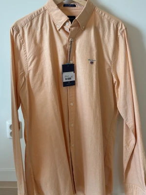 Beige långärmad skjorta från Gant - Snygg beige skjorta från Gant med klassisk button-down krage och broderad logga på bröstet. Skjortan har långa ärmar och knappar framtill. Perfekt för en clean och stilren look.